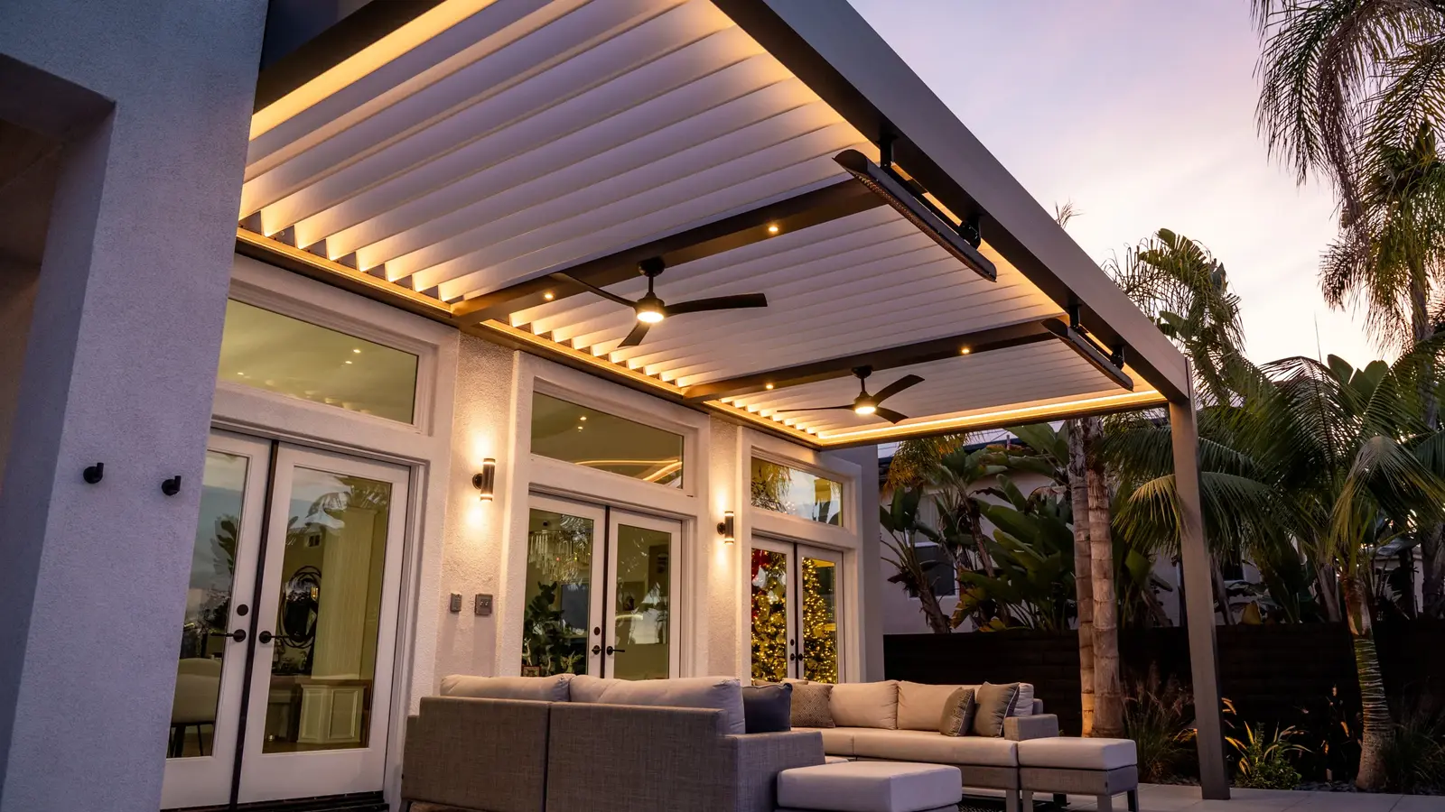 Two ceiling fans mounted to fixed beams beneath an illuminated Azenco R-BLADE louvered pergola