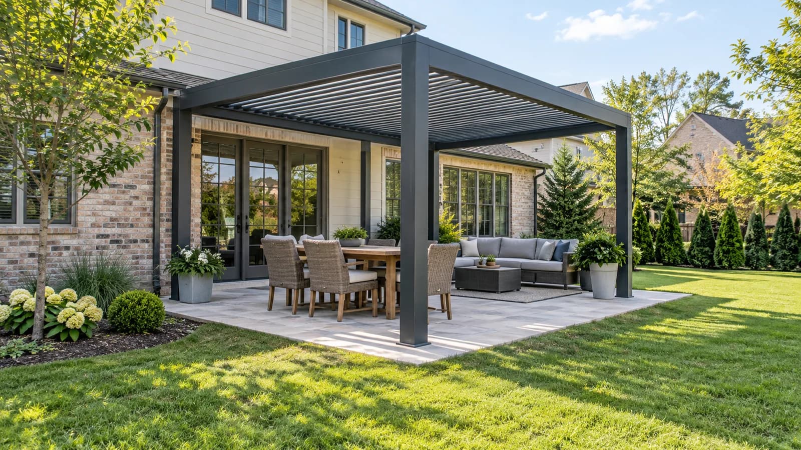 Charcoal aluminum louvered pergola over a light stone patio with open lawn beside a brick-and-siding home