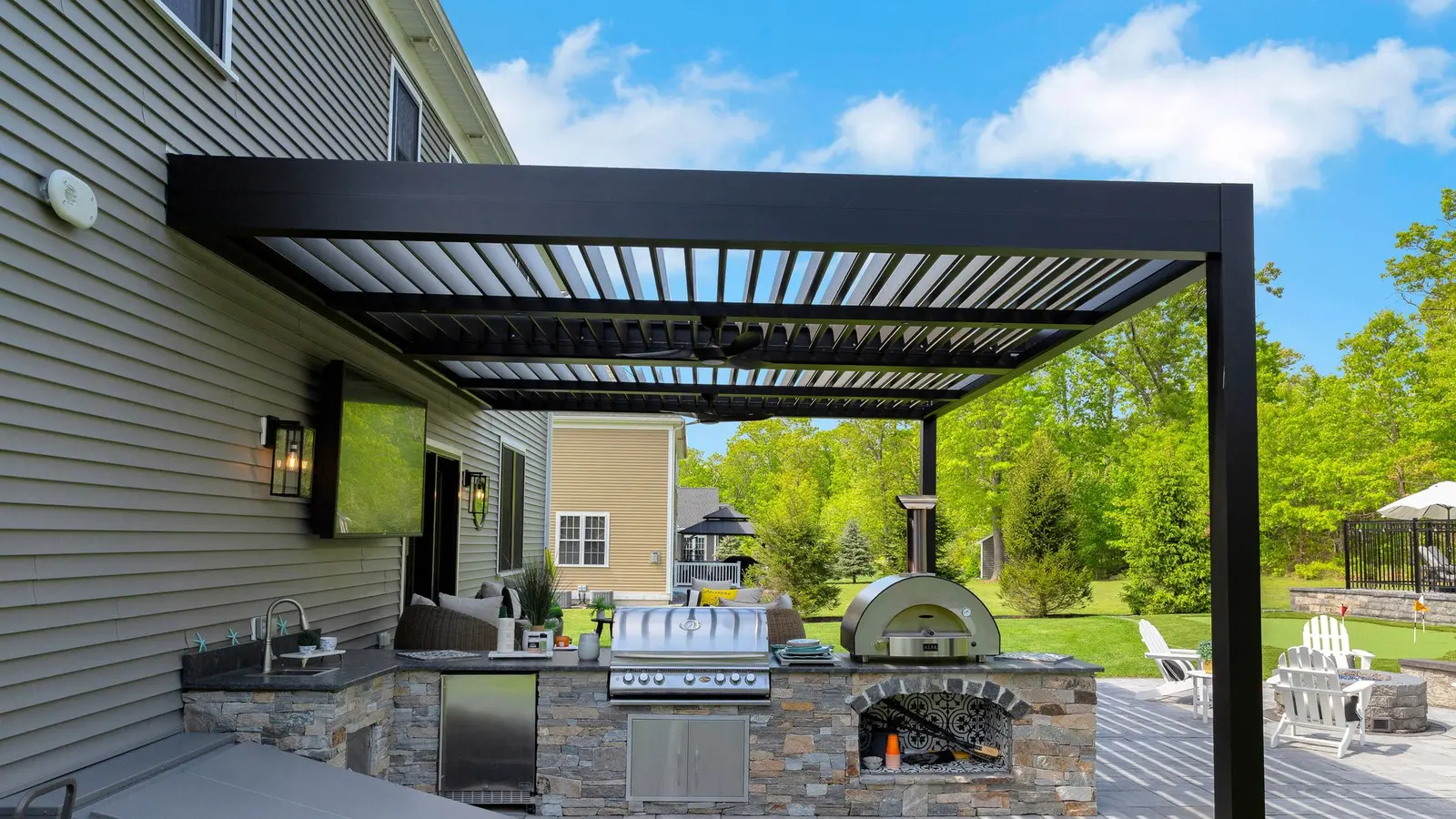 Built-in outdoor kitchen beneath a dark aluminum louvered pergola beside a home and patio seating